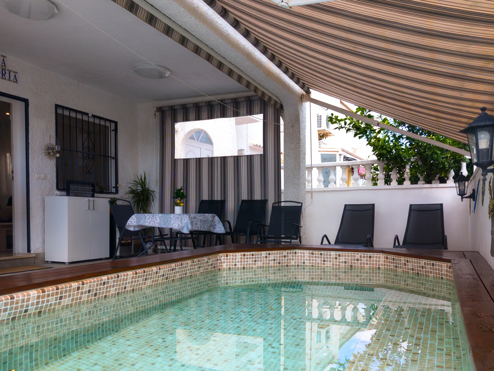 Small private pool with mosaic tiles in front of the covered terrace, with loungers, a table and chairs under striped awnings